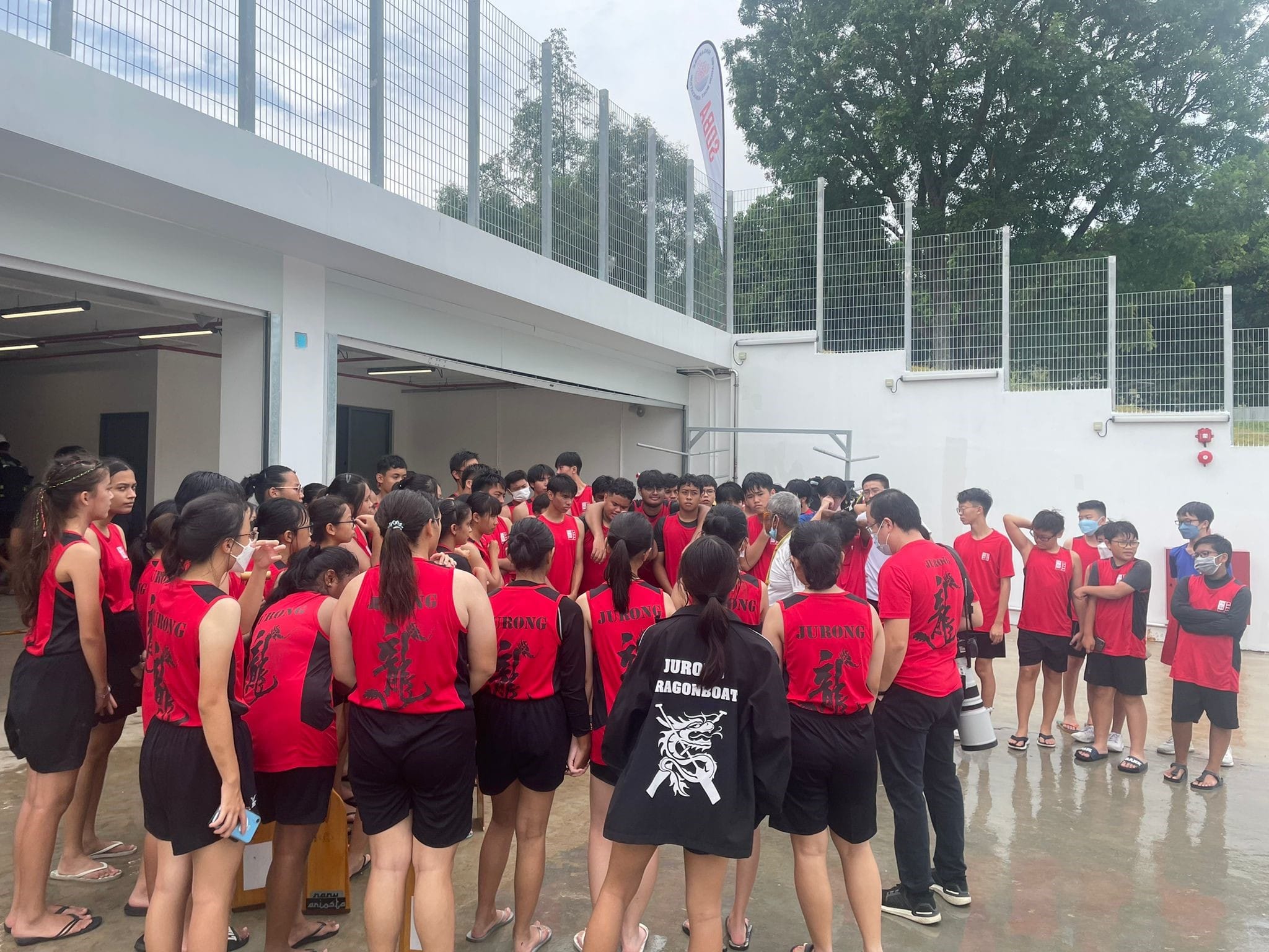 A large group of youth in red and black Jurong Dragon Boat uniforms gathered outdoors near a white building.